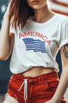 A female model wearing a unisex crew neck white t-shirt with hem and sleeves rolled up and the word America in red and an American flag in blue with red shorts