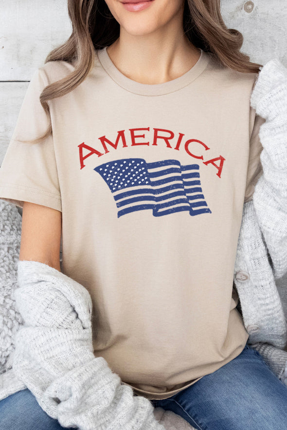 A female model wearing a unisex crew neck tan t-shirt and the word America in red and an American flag in blue with blue jeans