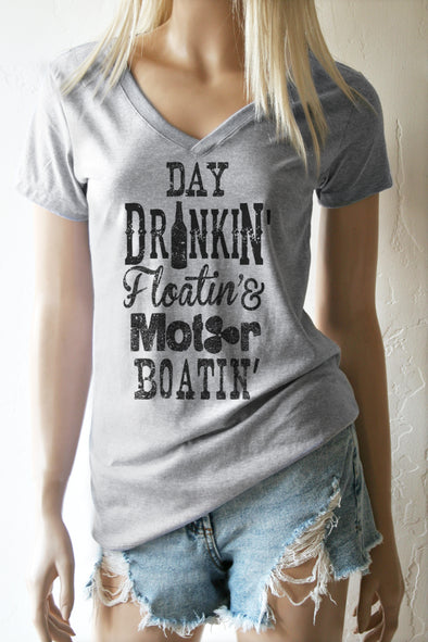 A blonde model standing wearing a heather grey v-neck t-shirt with the words in black Day Drinkin' Floatin' & Motor Boatin' and shredded cut off shorts