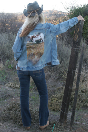 Woman standing outdoors with her hand on a fence post overlooking the pastures wearing a Longhorn Steer Denim Jacket. Jacket can be worn by Women or Men. Painting is on back of jacket.  - Southern Girl Apparel® - southerngirlapparel.com