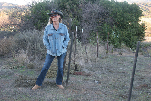 Woman standing outdoors with her back to the pastures looking at the camera, wearing a Denim Jacket with her hands in her pockets and a cowboy hat that has a bull on the front. Jacket can be worn by Women or Men. Painting of a Longhorn Steer is on the back of the denim jacket. - Southern Girl Apparel® - southerngirlapparel.com