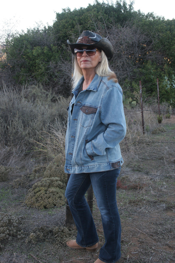 Woman standing sideways outdoors with her back to the pastures looking at the camera, wearing a Denim Jacket with her hands in her pockets and a cowboy hat that has a bull on the front. Jacket can be worn by Women or Men. Painting of a Longhorn Steer is on the back of the denim jacket. - Southern Girl Apparel® - southerngirlapparel.com
