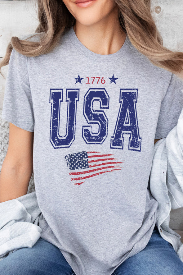 A female model wearing a heather grey short sleeve crew neck unisex t-shirt with 1776 in red at top and stars and USA in blue and red and blue American Flag below